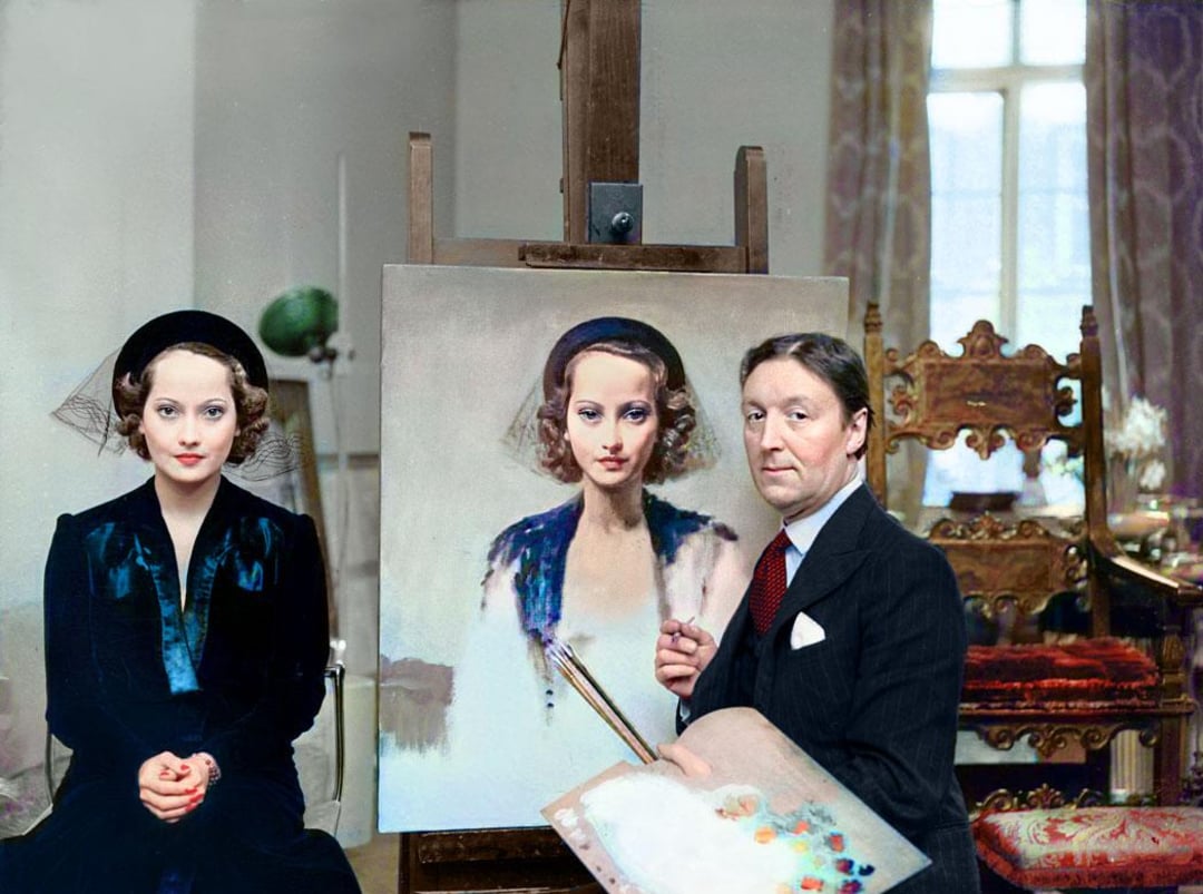 A woman in a dark outfit sits beside an artist, who is painting her portrait on a canvas. The artist holds a palette and brush, and they are in an ornate, well-lit room with large windows and elegant chairs.