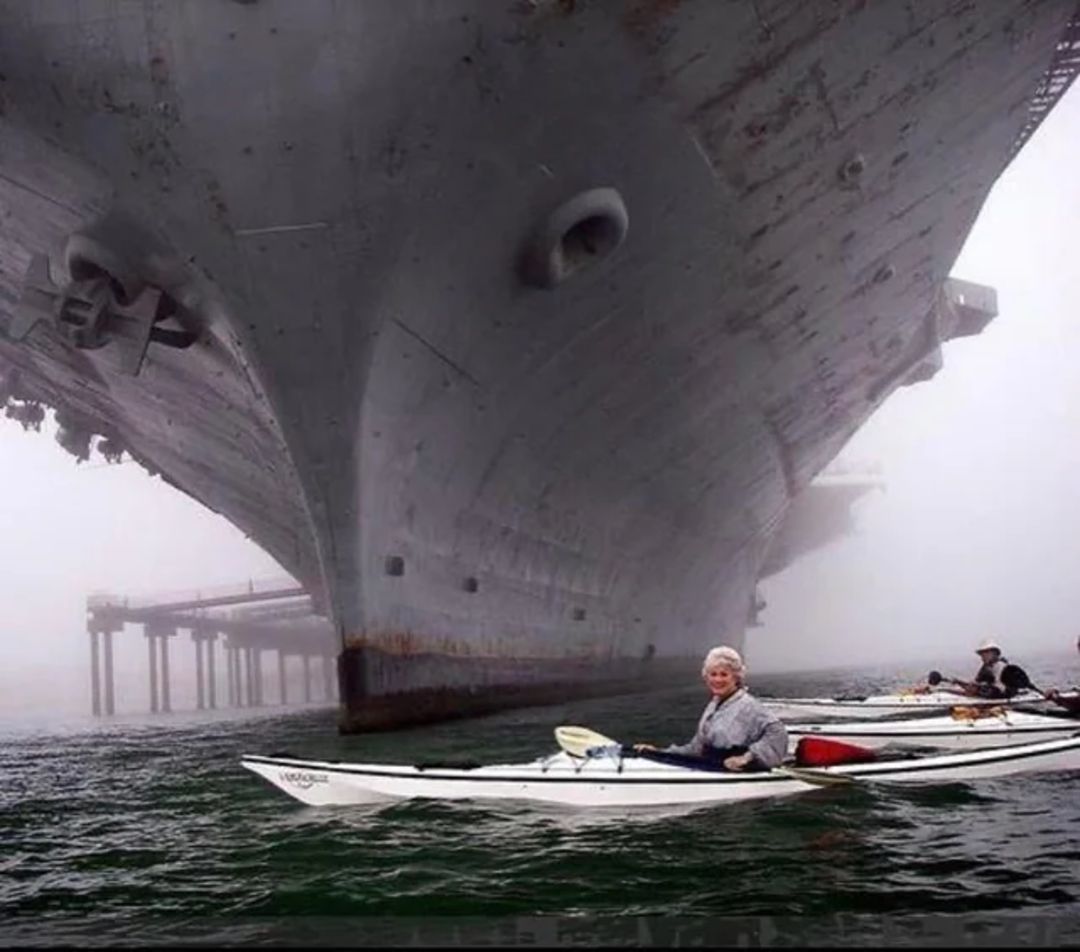 A group of people in kayaks paddle close to the bow of a massive ship, with fog obscuring the background and a dock partially visible to the side.
