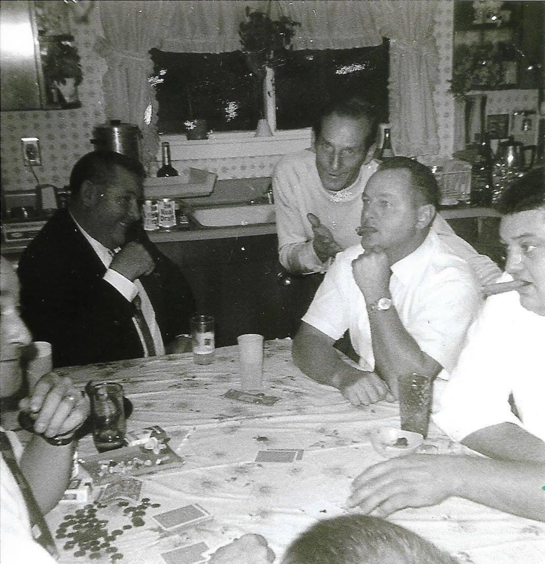 A group of men sit around a table playing cards, drinks and poker chips in front of them. One man stands behind, gesturing animatedly. The scene appears to be in a cozy, vintage kitchen.