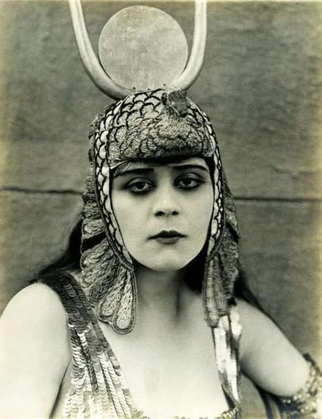 A woman in ancient Egyptian-style costume wears a headdress with a coiled cobra and large sun disk. Her expression is serious, and she has long dark hair and wears a garment with scale-like patterns. The image is in black and white.
