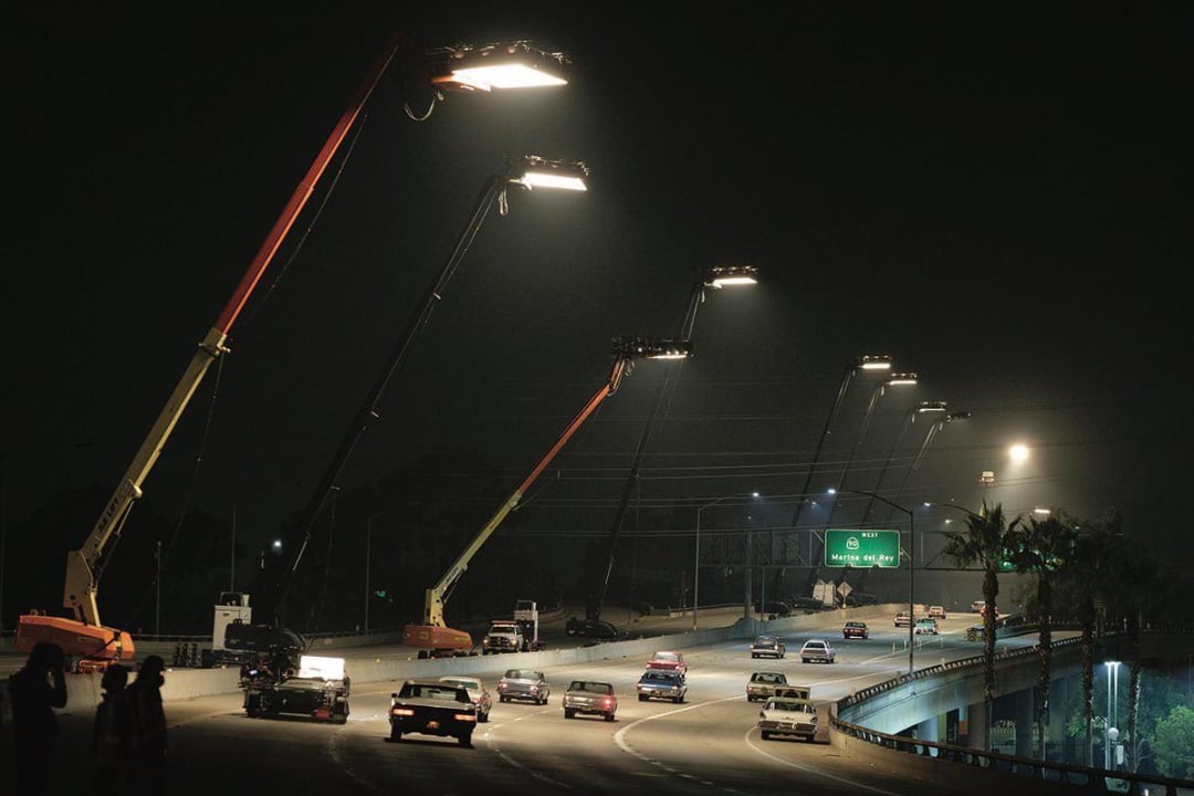 Several large cranes with bright lights are positioned over a busy highway at night, illuminating the road as cars drive below. Highway signs and palm trees are visible in the background.