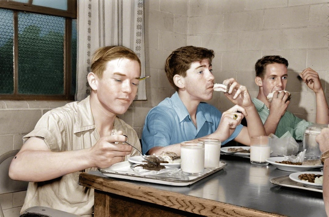 Three young men sit at a table in a cafeteria, eating meals and drinking glasses of milk. One holds a pencil behind his ear, while another takes a bite of bread. They appear to be engaged in quiet conversation.