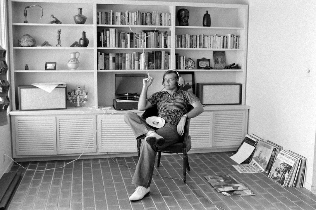 A man wearing headphones sits in a chair, holding a cigarette, in front of a shelf filled with books and objects. Vinyl records and a record player are nearby on the floor and a cabinet.