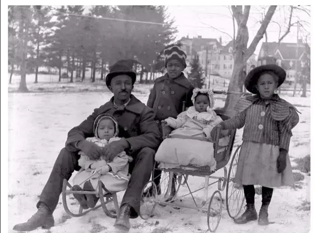 A Black man sits outdoors on a sled holding a young child, with three more children beside him. One child sits in a stroller, while the others stand. Snow covers the ground, and trees and buildings are in the background.