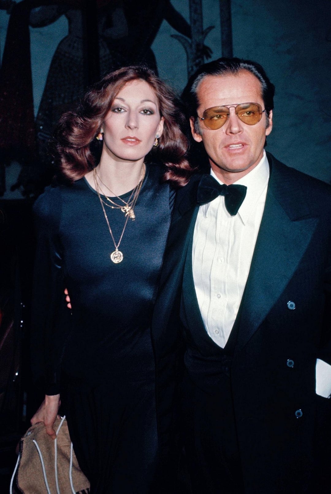 A woman in a black dress with layered necklaces stands beside a man in a tuxedo and tinted glasses; they both look at the camera while posing together at a formal event.