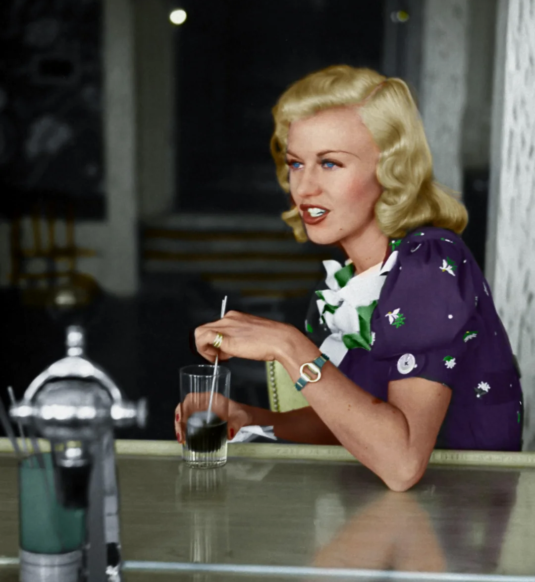 A woman with wavy blonde hair wearing a purple dress sits at a counter, stirring a drink with a straw and looking to the side, with a slight smile on her face.