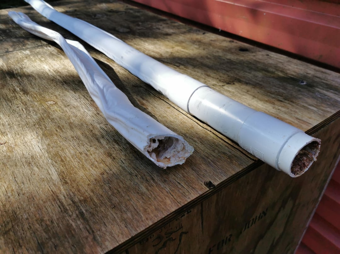 Two white PVC pipes with damaged, broken ends lie on a wooden surface outdoors, with one pipe more severely deteriorated than the other. Red siding is visible in the background.