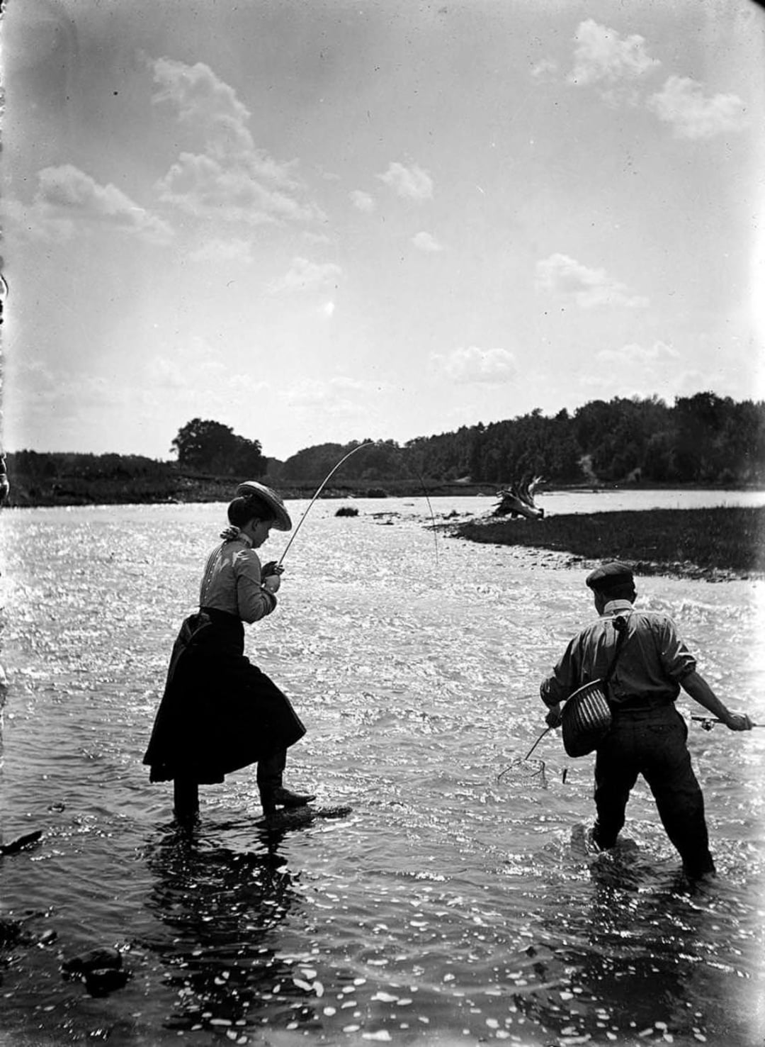A woman and a man stand knee-deep in a river while fishing. The woman holds a fishing rod, and the man carries a basket. Trees and a bright sky are visible in the background.