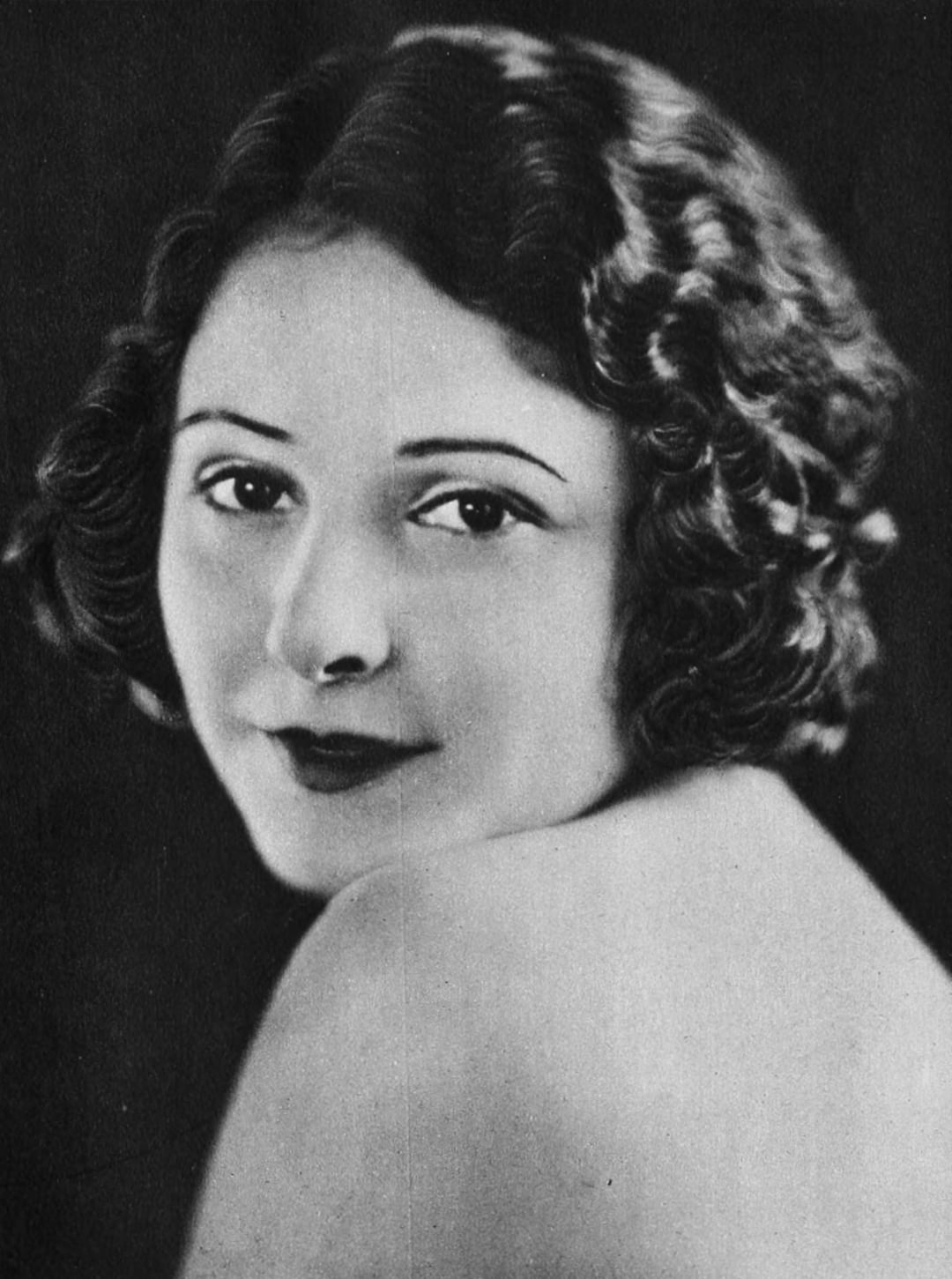 A woman with short, wavy dark hair gazes softly over her shoulder at the camera, with a neutral background behind her and a gentle expression on her face. The image is in black and white.
