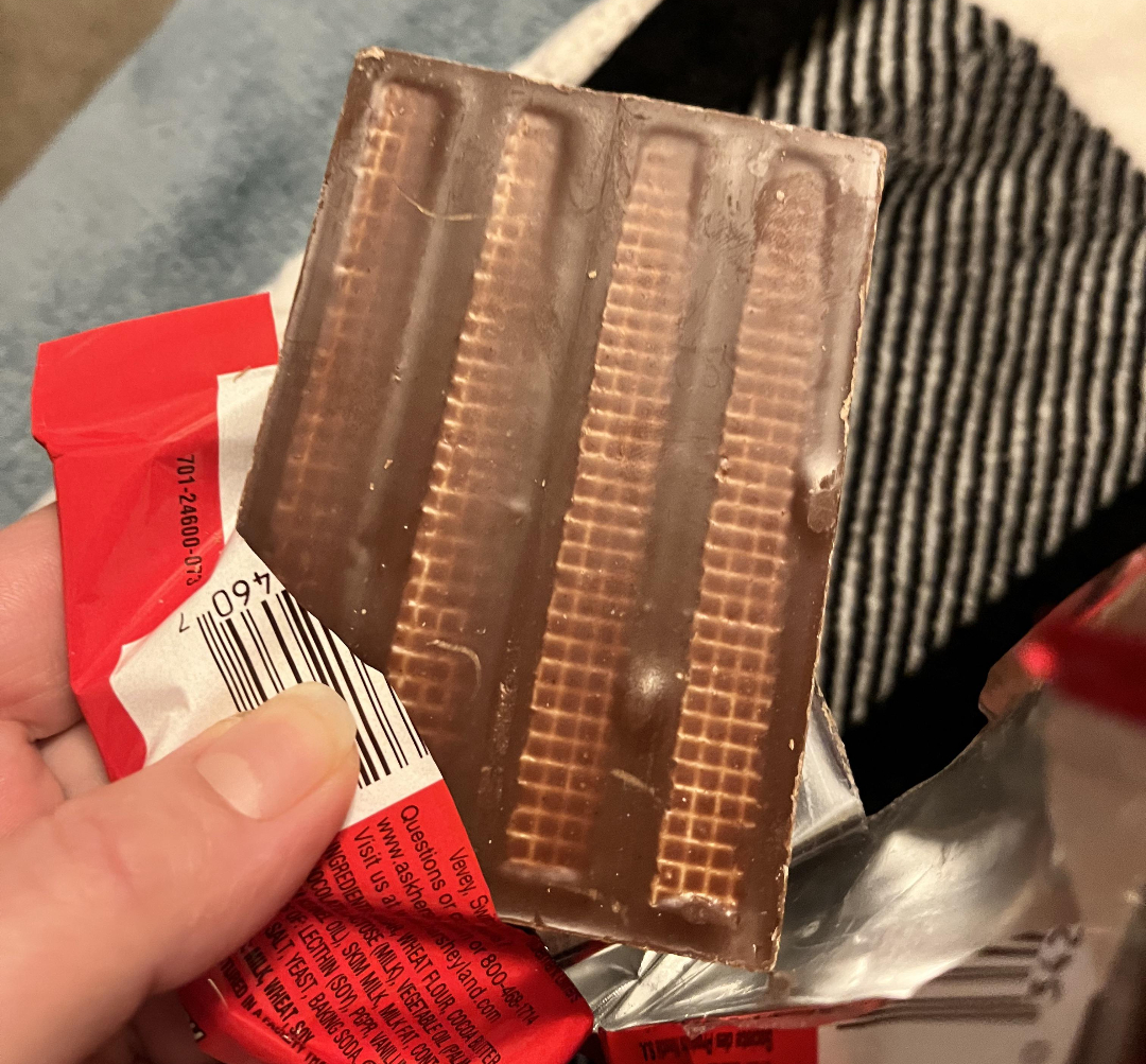 A hand holds a partially unwrapped chocolate bar, revealing wafer layers beneath the chocolate coating. The red and silver wrapper is crumpled around the bar.