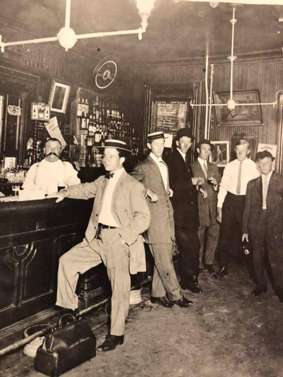 A vintage black-and-white photo shows several men in suits and hats standing at a bar with a bartender behind the counter. The bar is lined with bottles and glasses, and the decor appears to be from the early 1900s.