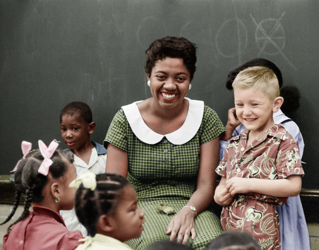 A smiling woman in a green dress sits in front of a chalkboard, surrounded by a diverse group of young children, some sitting and some standing. Everyone looks happy and engaged.