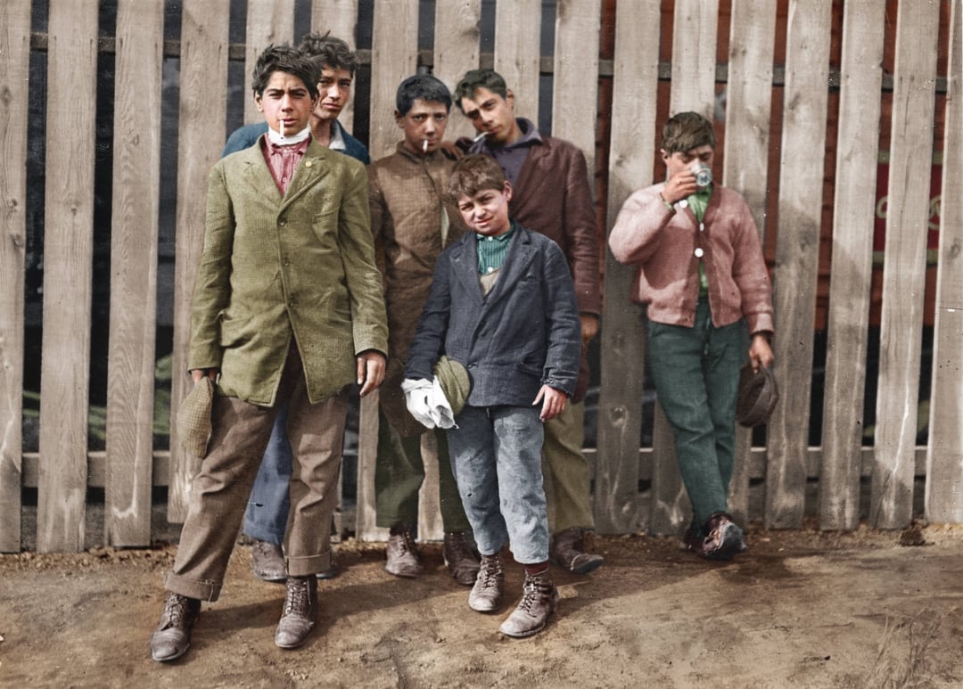 Six boys in early 20th-century clothing stand by a wooden fence; some have cigarettes in their mouths and wear worn jackets and boots, giving a sense of a historical, urban street scene.