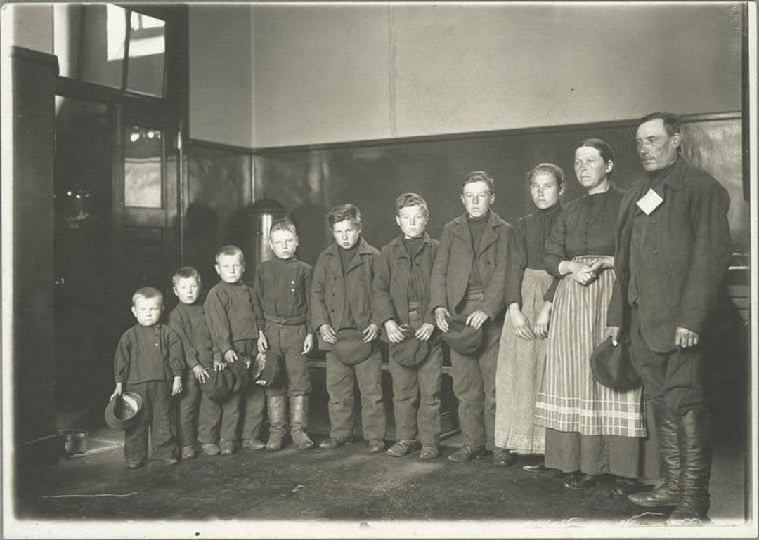 A black-and-white photo shows a family of ten, including eight children and two adults, standing in a line indoors. The children hold hats, and everyone wears plain, old-fashioned clothing.