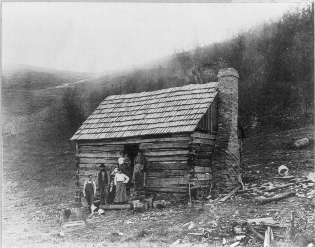 A black-and-white photo of five people standing outside a small log cabin with a stone chimney on a hillside. The area around the cabin is scattered with debris, logs, and household items. Sparse trees cover the hill behind.