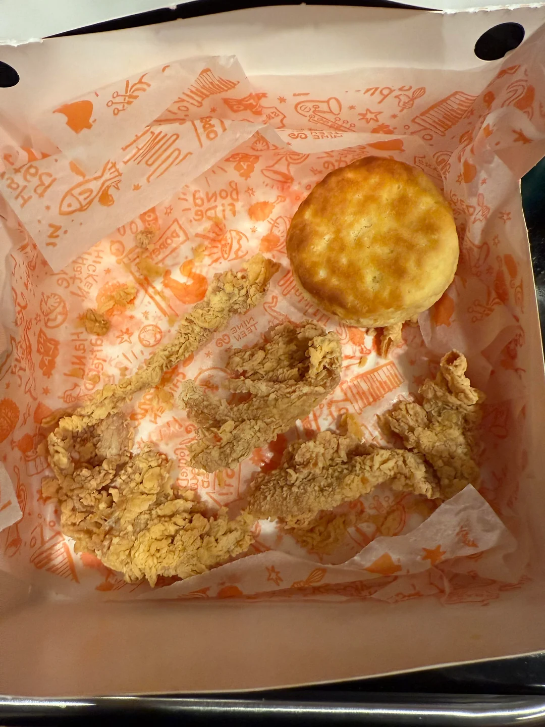 Open fast food box with a biscuit and three pieces of fried chicken on branded paper. The box appears mostly empty with leftover crumbs and grease marks.