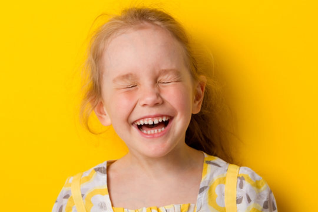 A young girl with light hair laughs with her eyes closed, standing in front of a bright yellow background. She is wearing a yellow and gray patterned dress.
