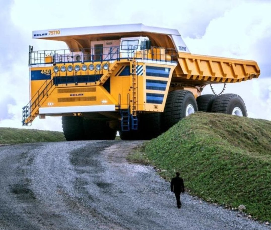 A person walks up a gravel road toward a massive yellow mining dump truck, which towers over the landscape with its large tires and elevated cab. The truck is labeled "BELAZ 75710.