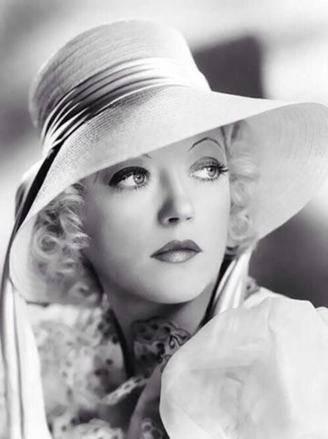 A woman with wavy blonde hair wears a large, elegant hat with a wide brim and ribbons. She has a thoughtful expression, dark lipstick, and is dressed in vintage-style clothing in a black and white portrait.