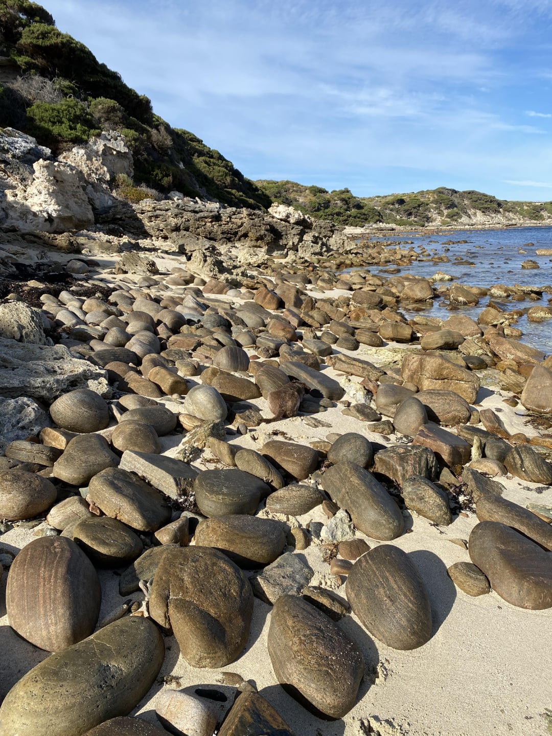 Large, smooth, rounded rocks and boulders scattered across a sandy beach with rocky cliffs and green vegetation in the background under a blue sky. The shoreline extends into the distance.