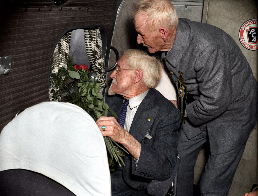 Two elderly men in suits sit inside an airplane. One holds a bouquet of roses and looks out the window, while the other leans over his shoulder, also gazing outside. The seat and airplane wall are visible.