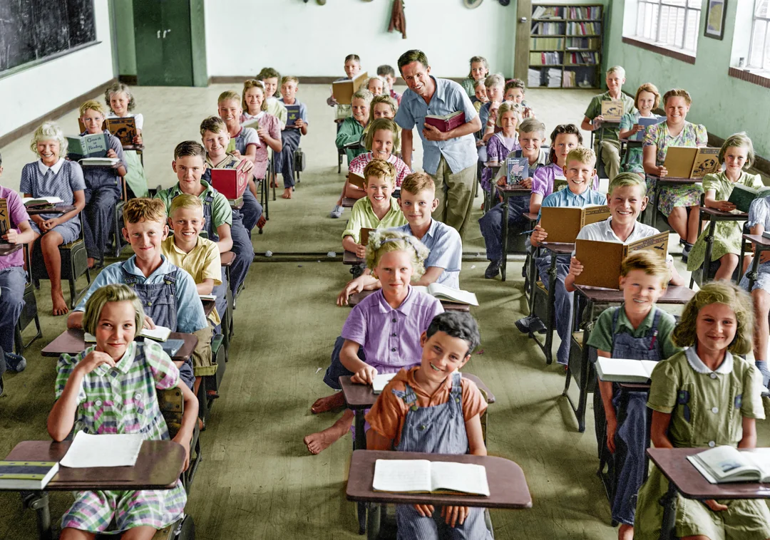 A vintage classroom filled with smiling children seated at desks, holding books and notebooks, with two adults standing in the back. Some students are barefoot and the room has large windows and bookshelves.