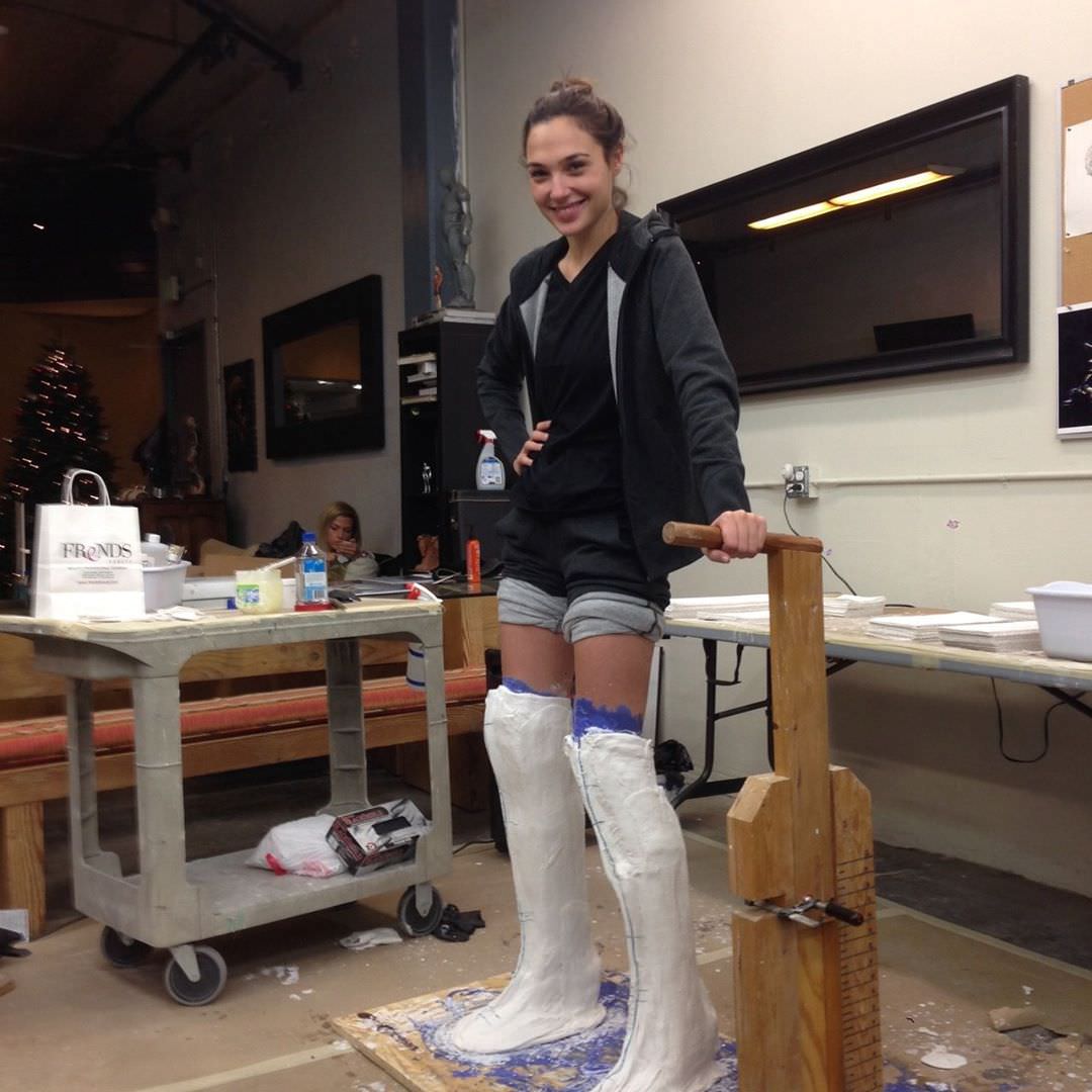 A smiling woman in a gray hoodie and shorts stands in a workshop with her legs covered in white plaster casting material, holding a wooden support frame. Work tables and supplies are visible in the background.