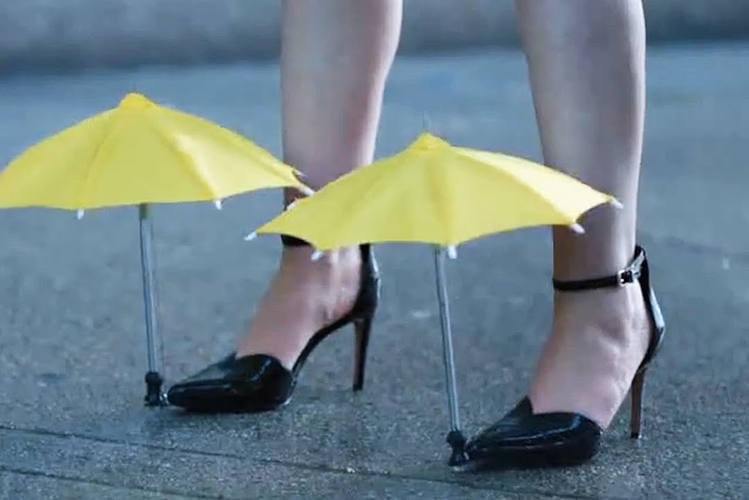 A person wearing black high heels stands on wet pavement, with small yellow umbrellas attached above each shoe to shield them from rain.