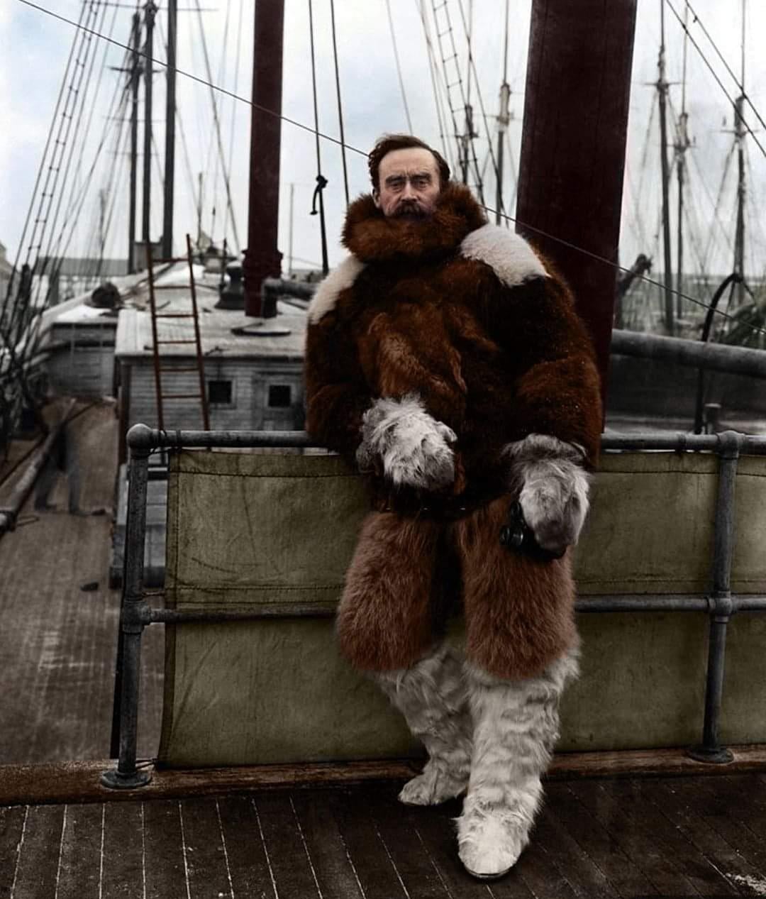 A man with a brown beard, dressed in a thick fur coat and pants, stands on the deck of a wooden ship with tall masts and rigging visible in the background.
