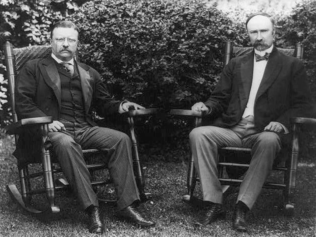 Two men in formal suits sit on wooden rocking chairs outdoors, with dense leafy bushes behind them. Both have mustaches and serious expressions, and the photo appears to be from the early 1900s.