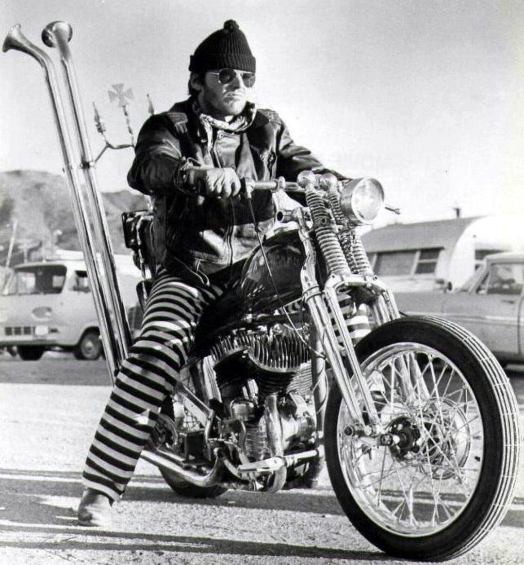 A person wearing a beanie, sunglasses, striped pants, and a leather jacket sits on a custom motorcycle with tall exhaust pipes, parked in a lot with cars and trailers in the background.
