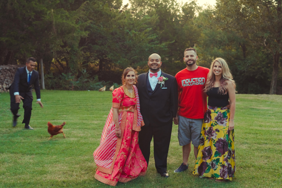 Four people pose for a photo on a grassy lawn; one wears a suit, one a red T-shirt, one a pink sari, and one a floral skirt. In the background, a man chases a chicken across the grass near some trees.