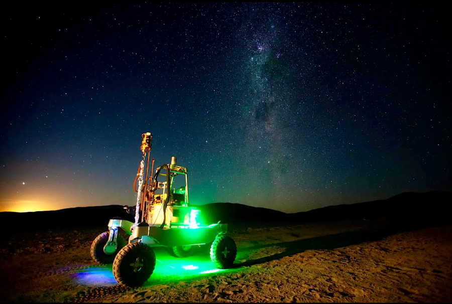 A small robotic rover with glowing green lights stands on desert terrain at night under a clear, star-filled sky and the visible Milky Way. Dark hills are visible on the horizon.