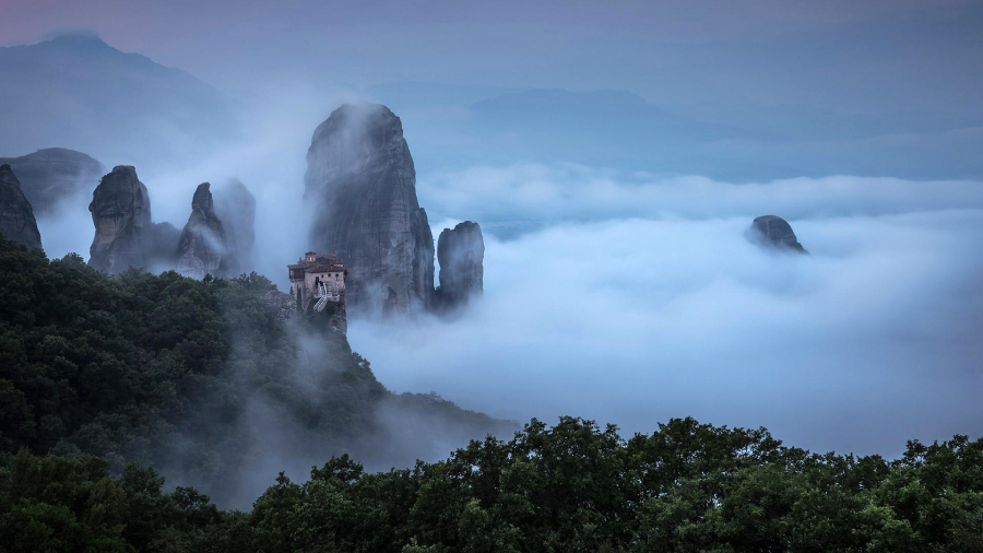 A monastery sits atop a rocky cliff surrounded by dense mist and towering rock formations, with lush green trees in the foreground and a mountain range fading into the distance.