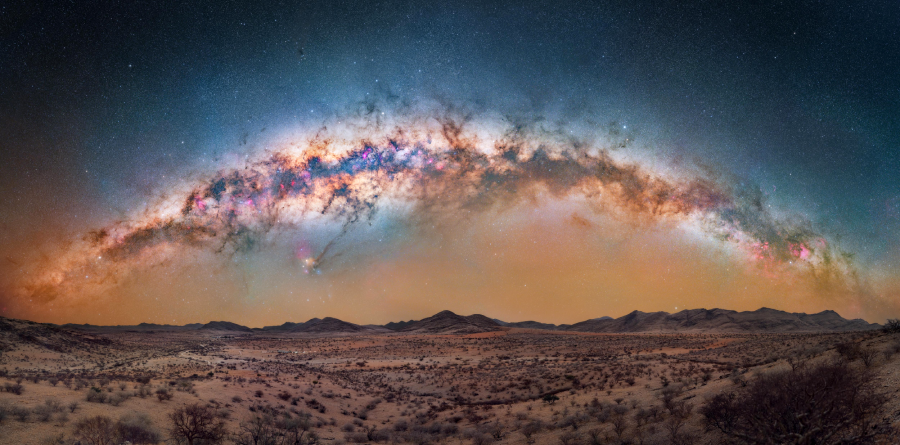 A panoramic view of a desert landscape under a clear night sky, with the vibrant, colorful Milky Way galaxy arcing across the horizon above distant hills.