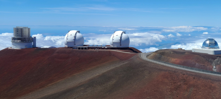Observatories with large white domes sit atop a barren, reddish-brown mountain above the clouds under a bright blue sky. A winding road leads to the domes, which are surrounded by a dramatic high-altitude landscape.