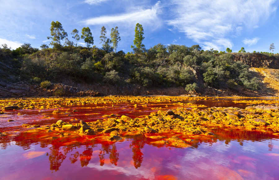 A river with striking red-orange water flows through a rocky landscape, with green trees and bushes on the opposite bank under a blue sky with wispy clouds.
