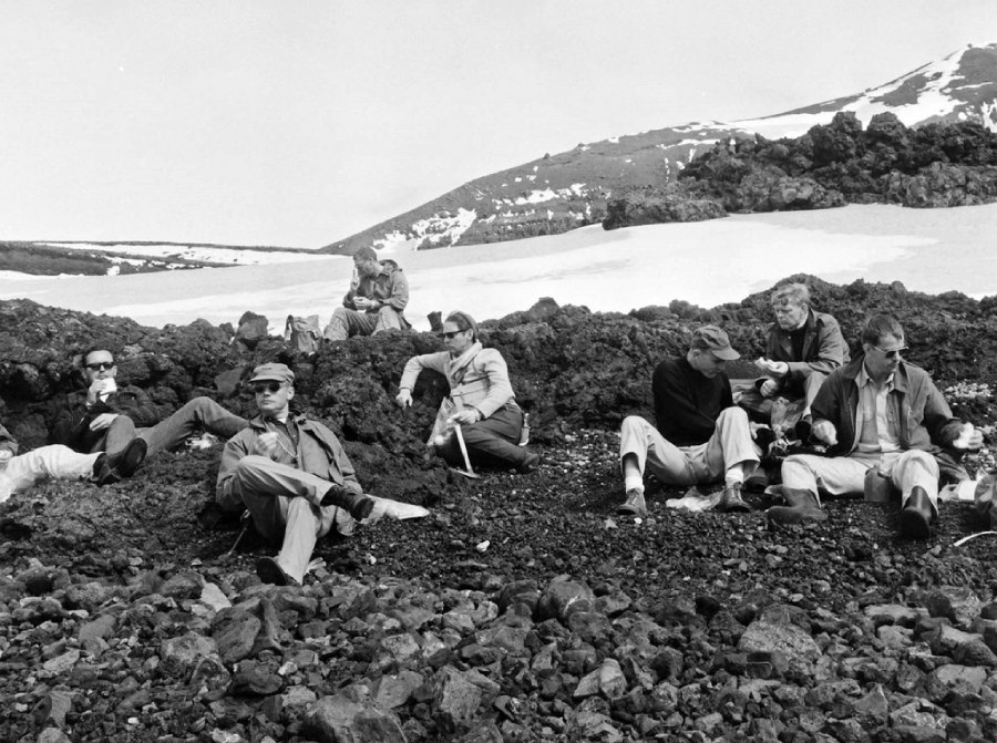 A group of men sit and relax on rocky ground with snow patches in the background, possibly on a mountain or volcanic terrain, dressed in outdoor clothing and sunglasses, appearing to rest or have a discussion.