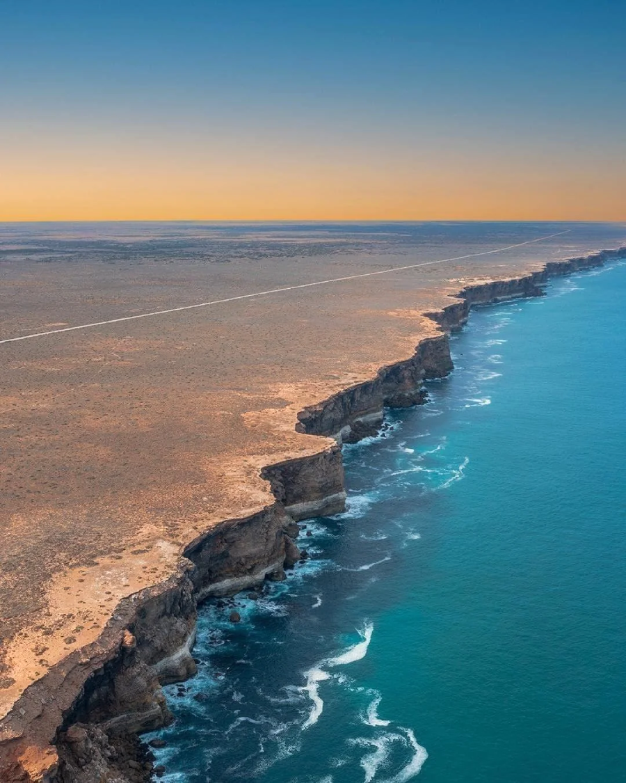 Aerial view of dramatic sea cliffs meeting turquoise ocean waves under a clear sky, with a flat, barren landscape stretching inland and a straight road visible near the cliffs’ edge.