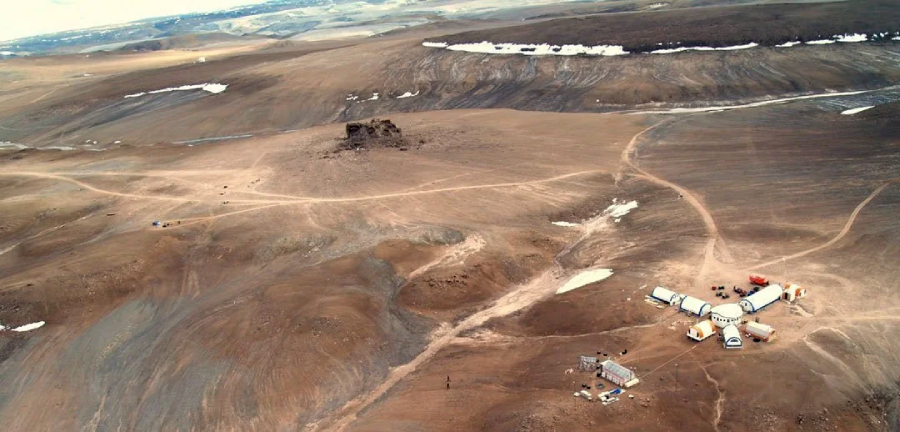 A cluster of small buildings and tents sits on a barren, rocky landscape with distant hills and patches of snow under a cloudy sky, suggesting a remote research camp in an arid, polar region.