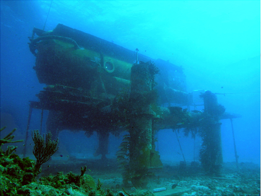 A large underwater research habitat stands on stilts on the ocean floor, surrounded by marine life and blue water, with algae and corals growing on its metal structure.
