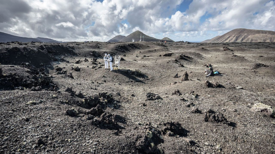 People in white spacesuits stand on a rocky, barren landscape with volcanic hills and mountains in the background under a partly cloudy sky, simulating an extraterrestrial environment.