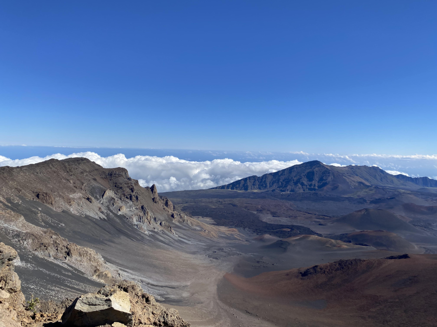 A wide volcanic crater with rocky slopes and sparse vegetation under a clear blue sky. Clouds float below the mountain peaks in the background, giving a sense of high elevation and open landscape.