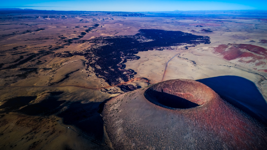 Aerial view of a volcanic crater with a dark lava flow spreading across a vast, arid landscape under a clear blue sky. Shadows highlight the crater's rim and surrounding terrain.