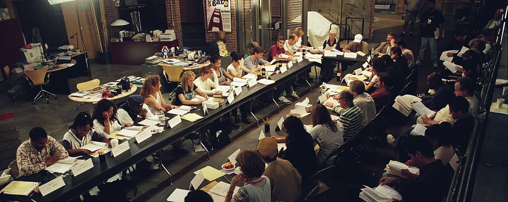 A large group of people sit around two long tables arranged in a rectangle, reading scripts and papers in what appears to be a rehearsal or meeting space with various office and studio supplies in the background.