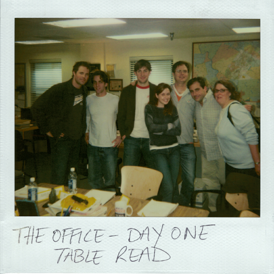 A group of seven people stand close together and smile inside an office, next to desks with papers and drinks. Handwritten text below reads: "THE OFFICE - DAY ONE TABLE READ.