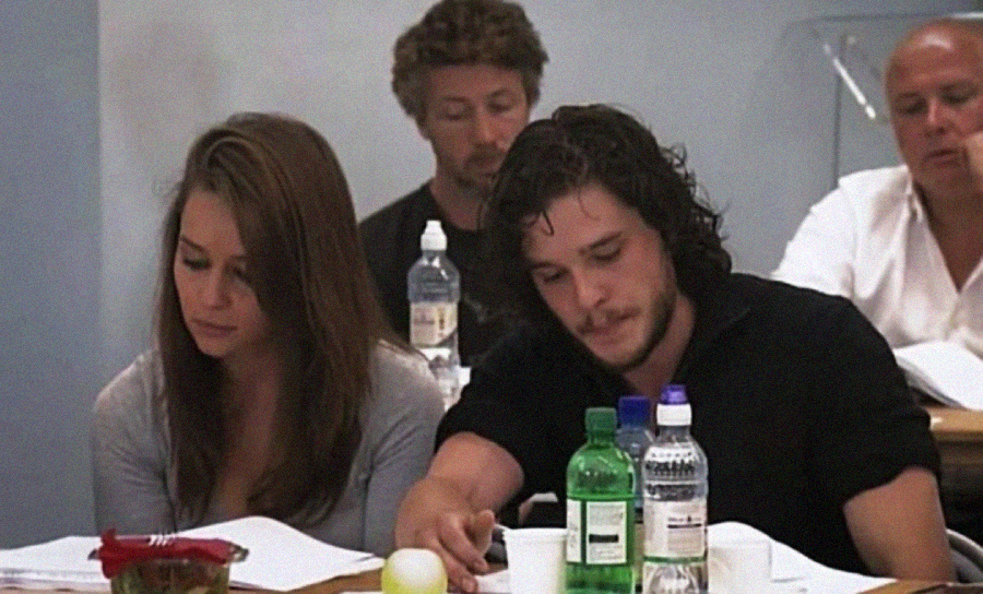 A group of people sit at a table reading scripts, with water bottles and snacks in front of them. Two individuals in the foreground appear focused on their papers.