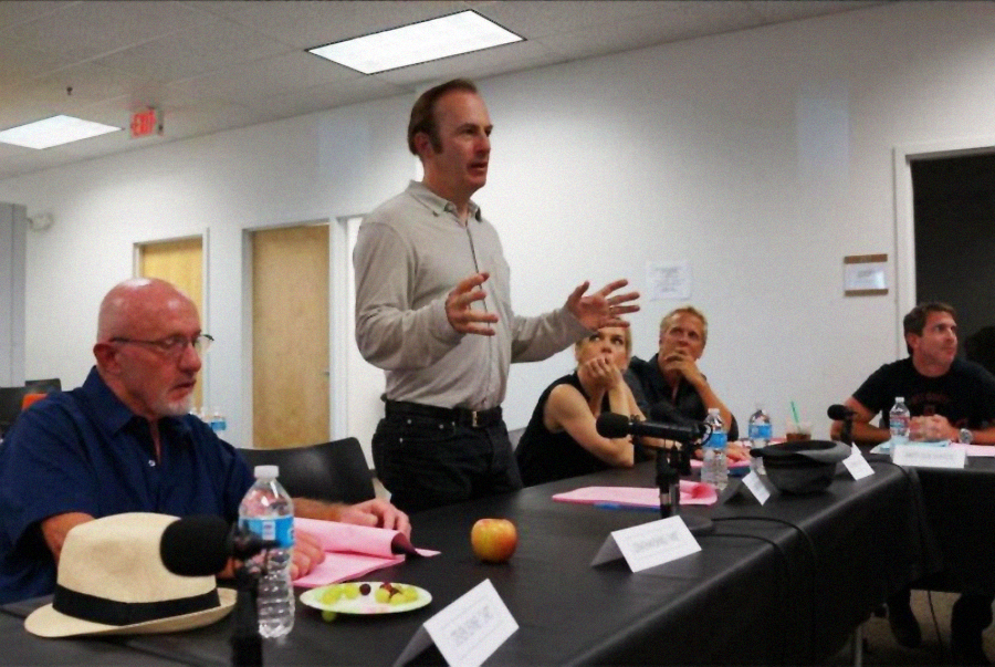 A man stands and speaks while others sit around a conference table covered with scripts, water bottles, and snacks, suggesting a meeting or script reading in a casual office setting.