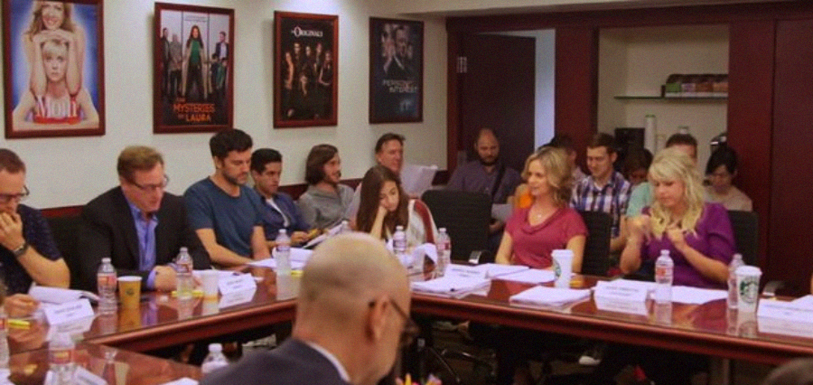 A group of people sit around a long table at a script reading session, with scripts, water bottles, and coffee cups in front of them. Posters of TV shows decorate the walls in the background.