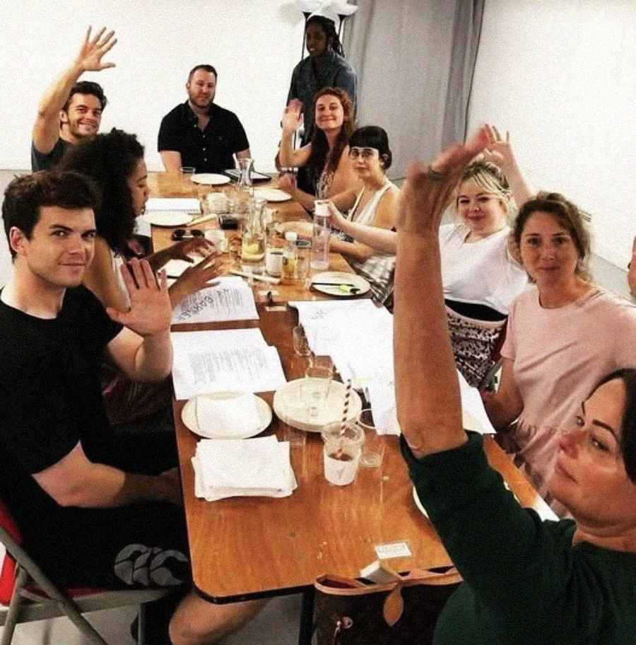 A group of people sit around a long table covered with papers, plates, and glasses, smiling and waving at the camera in a well-lit room.
